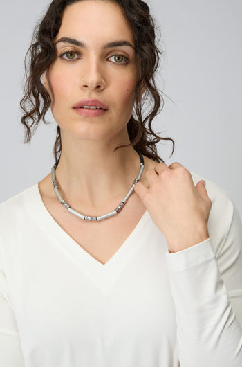 Woman with wavy dark hair in a white V-neck top wears the Silver Aluminium Hematite Necklace by Christina Brampti, gazing at the camera as her hand touches the striking accessory.