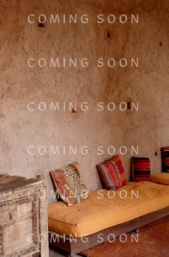 A rustic room with a textured plaster wall, wooden cabinet, and low seating with patterned cushions—perfect for lounging in the Cotton Jersey Boxy Pocket Top.