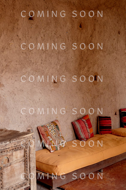 A rustic room with a textured plaster wall, wooden cabinet, and low seating with patterned cushions—perfect for lounging in the Cotton Jersey Boxy Pocket Top.