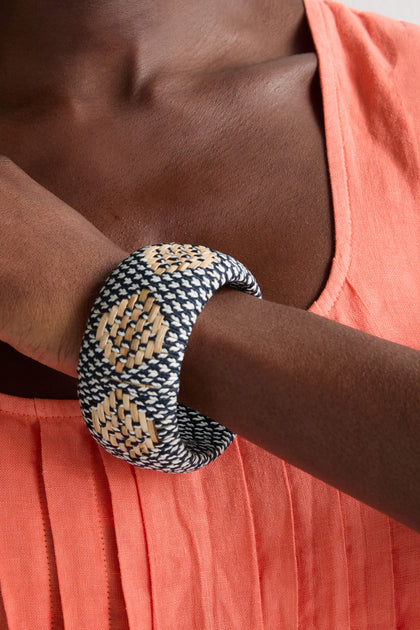 A person wearing a coral pleated top and a Handwoven Rattan Detail Bangle, a statement accessory with black, beige, and white patterns, on their wrist.