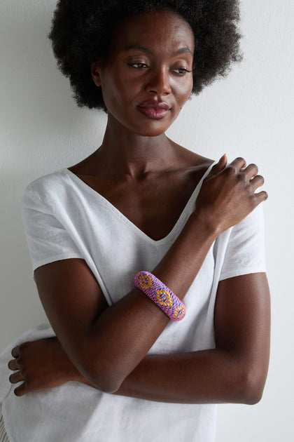 A woman in a white V-neck shirt poses with crossed arms, highlighting the Handwoven Fluoro Bangle—expertly crafted using traditional methods and vibrant neon tones—set against a plain light background.