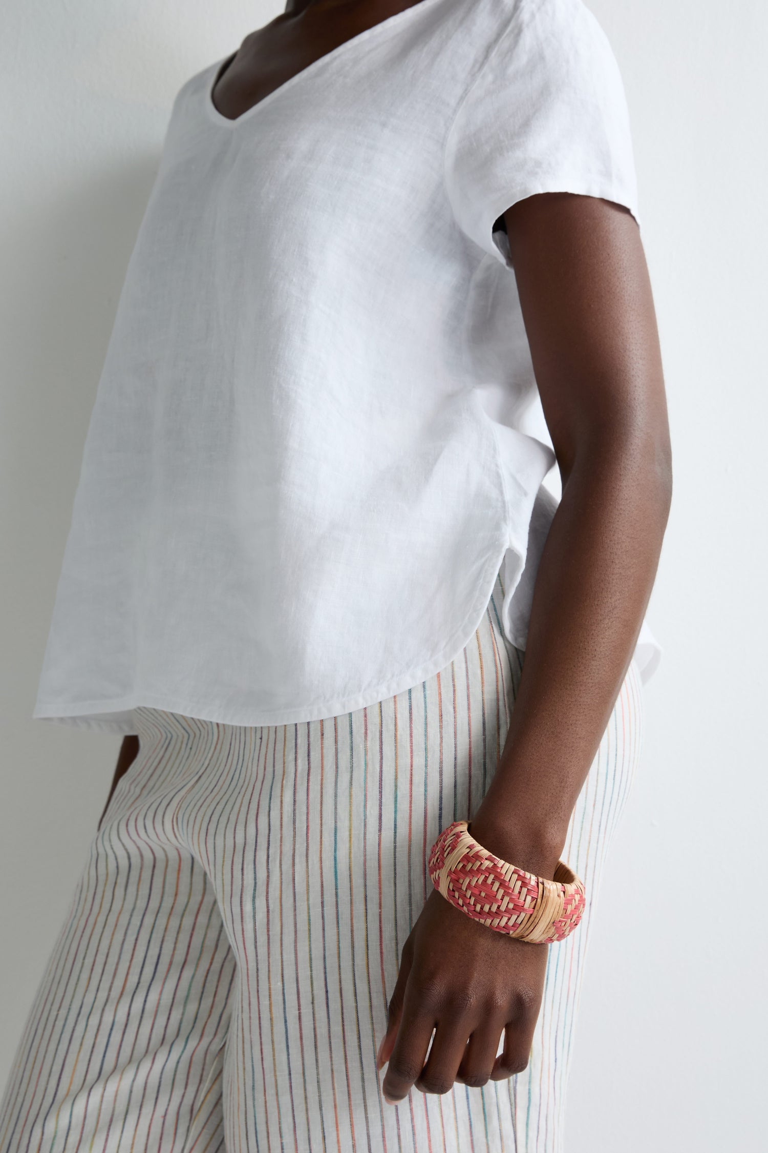 A person stands against a plain white wall wearing a loose white short-sleeve top, striped light-colored pants, and the Handwoven Rose Rattan Bangle, celebrating artisanal craftsmanship.