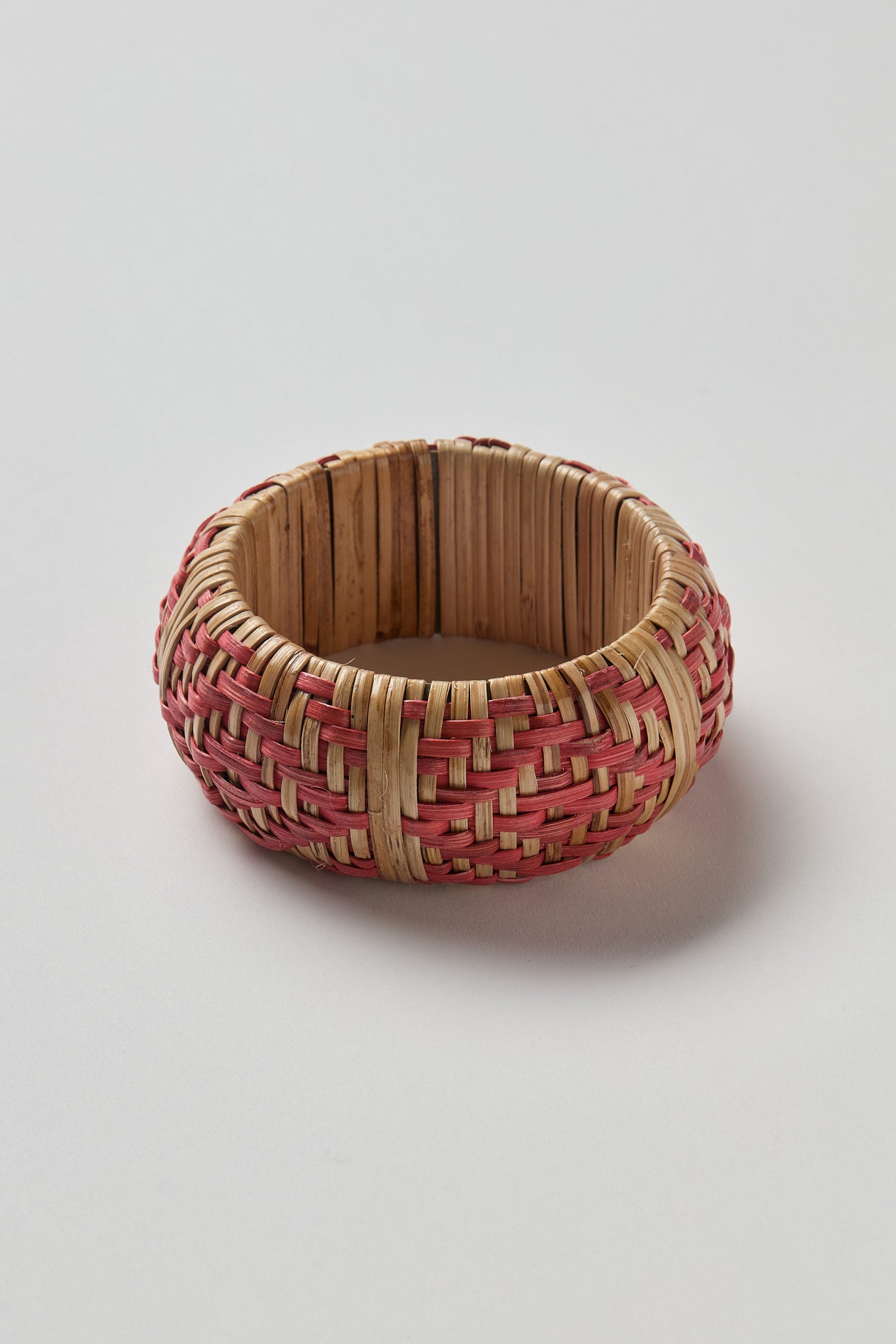 The Handwoven Rose Rattan Bangle features round, natural and pink-toned rattan, expertly woven to highlight artisanal tradition and natural beauty, presented against a plain white background.