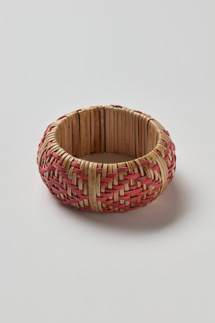 The Handwoven Rose Rattan Bangle features round, natural and pink-toned rattan, expertly woven to highlight artisanal tradition and natural beauty, presented against a plain white background.