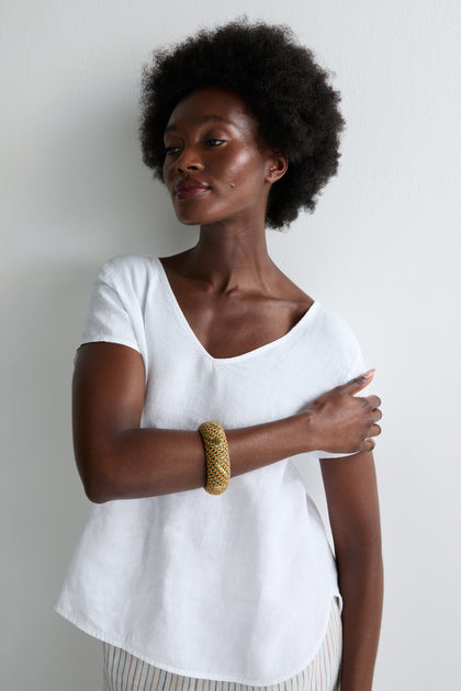 A woman with short, curly hair in a white shirt wears the Handwoven Multicoloured Bangle, known for its vibrant threads and heritage craftsmanship, as she stands against a white wall with one arm across her body.