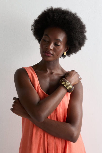 A woman with natural hair in a sleeveless coral dress stands against a white background, arms crossed and looking down. She completes her look with gold earrings and the Handwoven Espresso Bangle, detailed with golden threads.