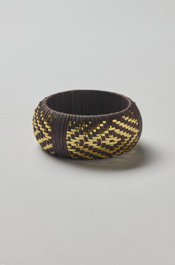 The Handwoven Espresso Bangle features a round shape with a yellow and dark brown zigzag pattern, crafted with golden threads, and is shown on a plain light background.