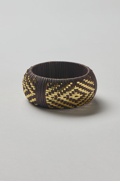 The Handwoven Espresso Bangle features a round shape with a yellow and dark brown zigzag pattern, crafted with golden threads, and is shown on a plain light background.