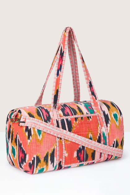 A colorful Ikat Velvet Weekend Bag featuring a multicolored abstract print, two handles, and an adjustable shoulder strap against a plain background highlights the traditional resist-dyeing technique used in its creation.