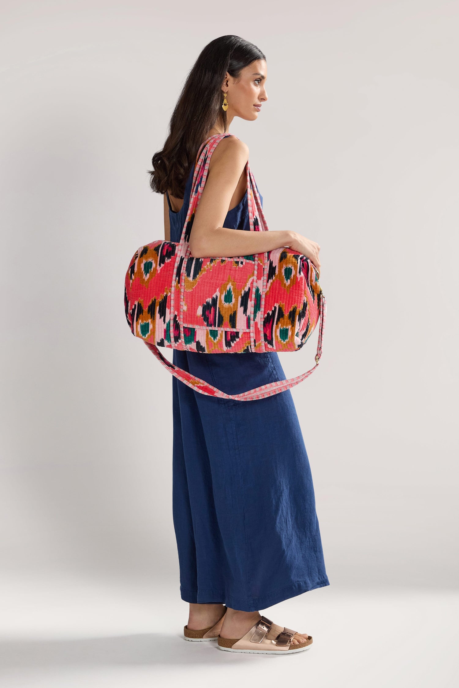 A woman in a blue dress and sandals poses with an Ikat Velvet Weekend Bag, showcasing its versatile design against a plain background.