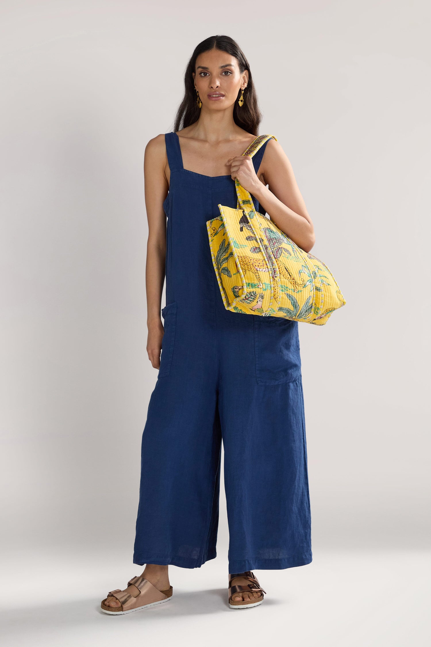 A woman stands wearing a dark blue jumpsuit and brown sandals, holding a Jungle Print Velvet Bag.