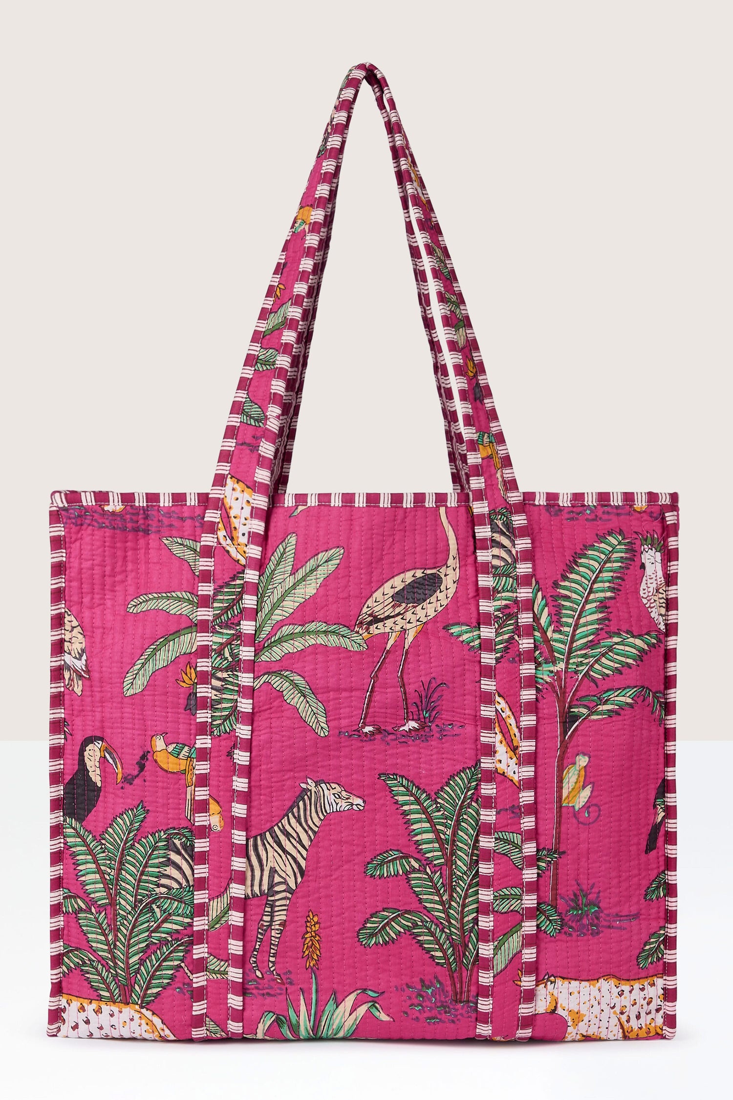 A Jungle Print Tote Bag from Conscious Yoga Collective features a colorful array of animals and plants, including zebras, birds, and palm leaves. The bag boasts long striped handles and a quilted fabric texture.