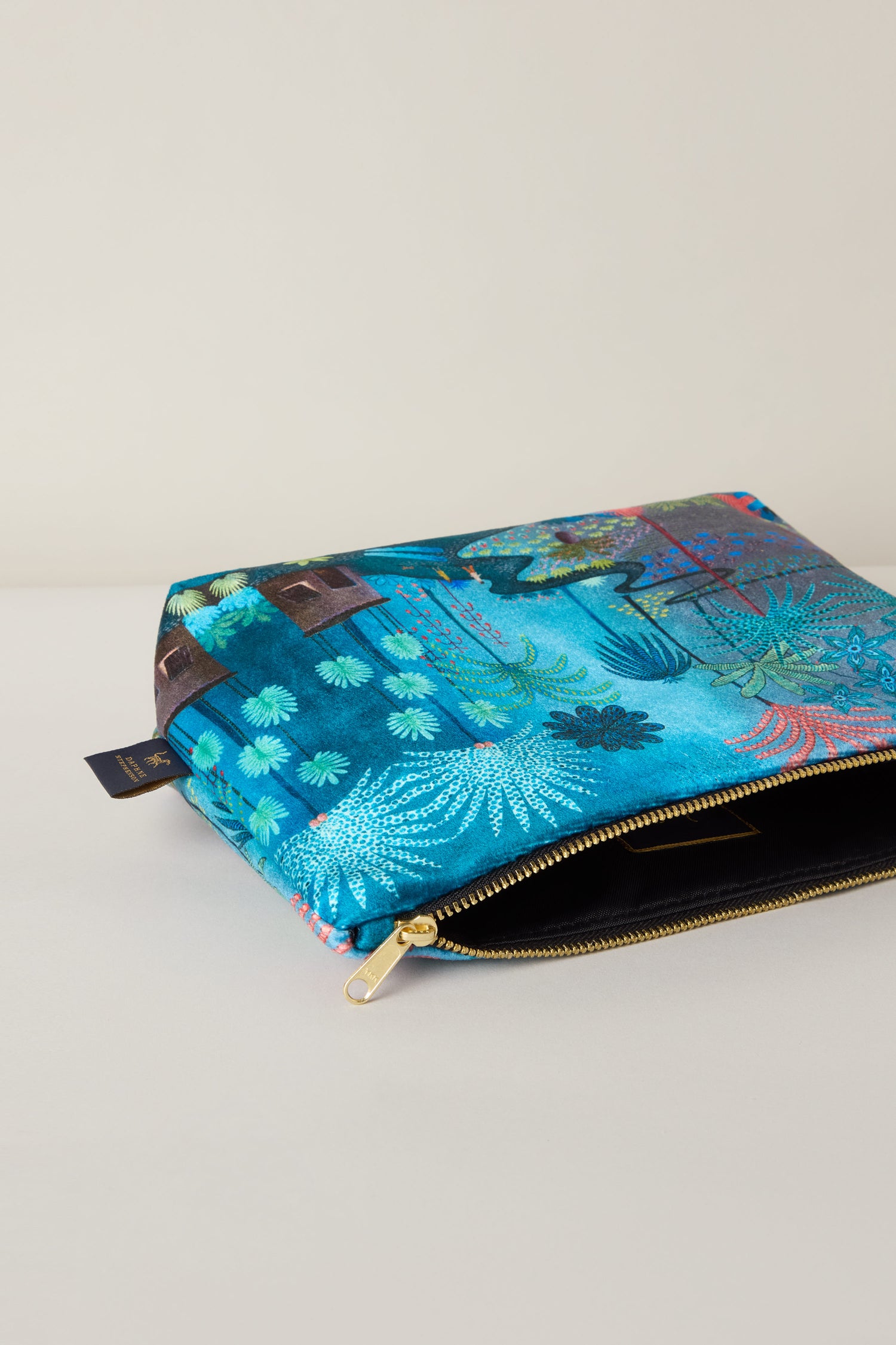 The Large Jungle Pathway Velvet Bag, created by Daphne Stephenson, sits open on a flat surface. Its blue zippered exterior displays a lively pattern of trees and plants.