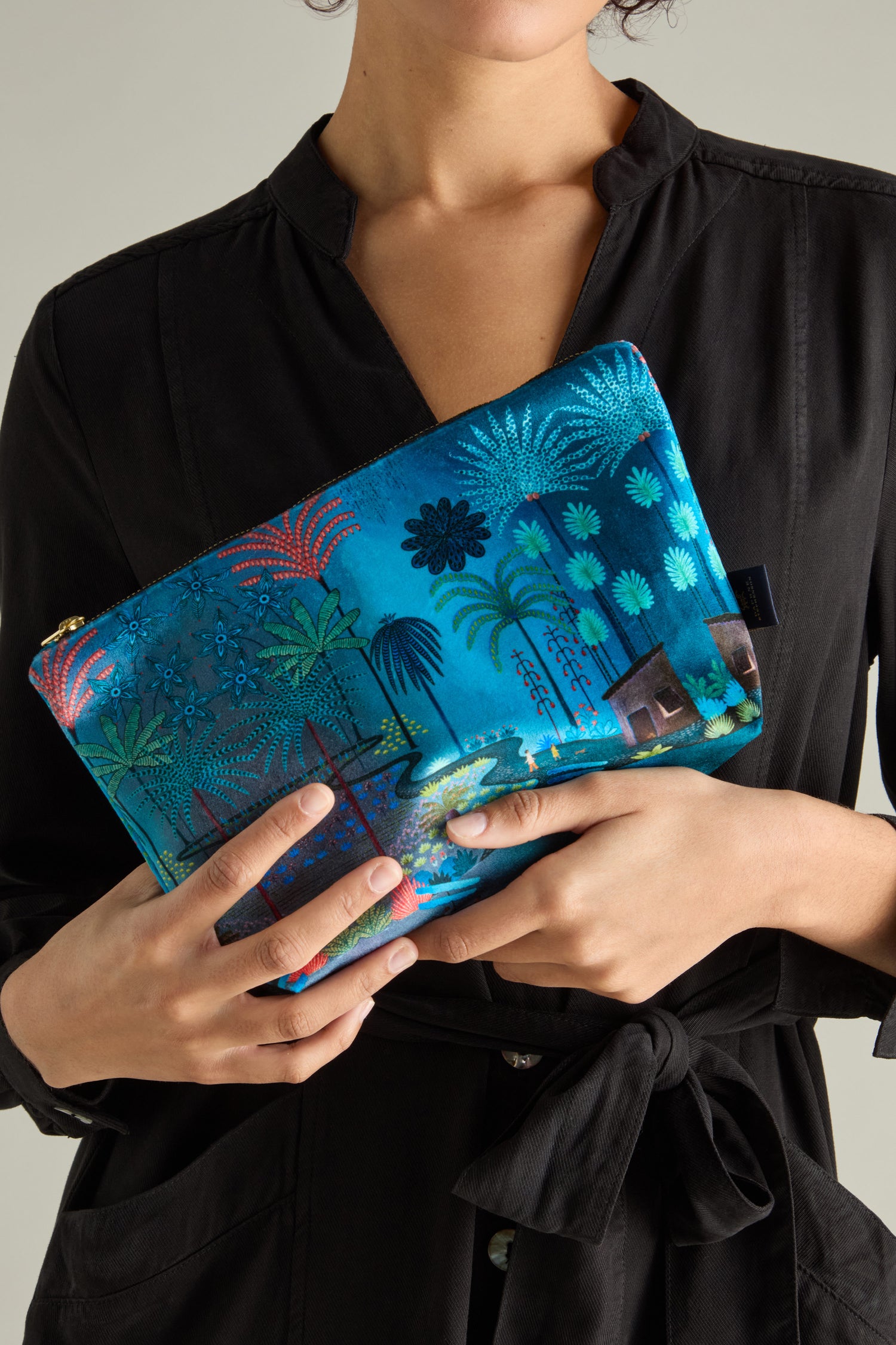 A person in a black dress holds a Large Jungle Pathway Velvet Bag by Daphne Stephenson, featuring vibrant palm tree designs.