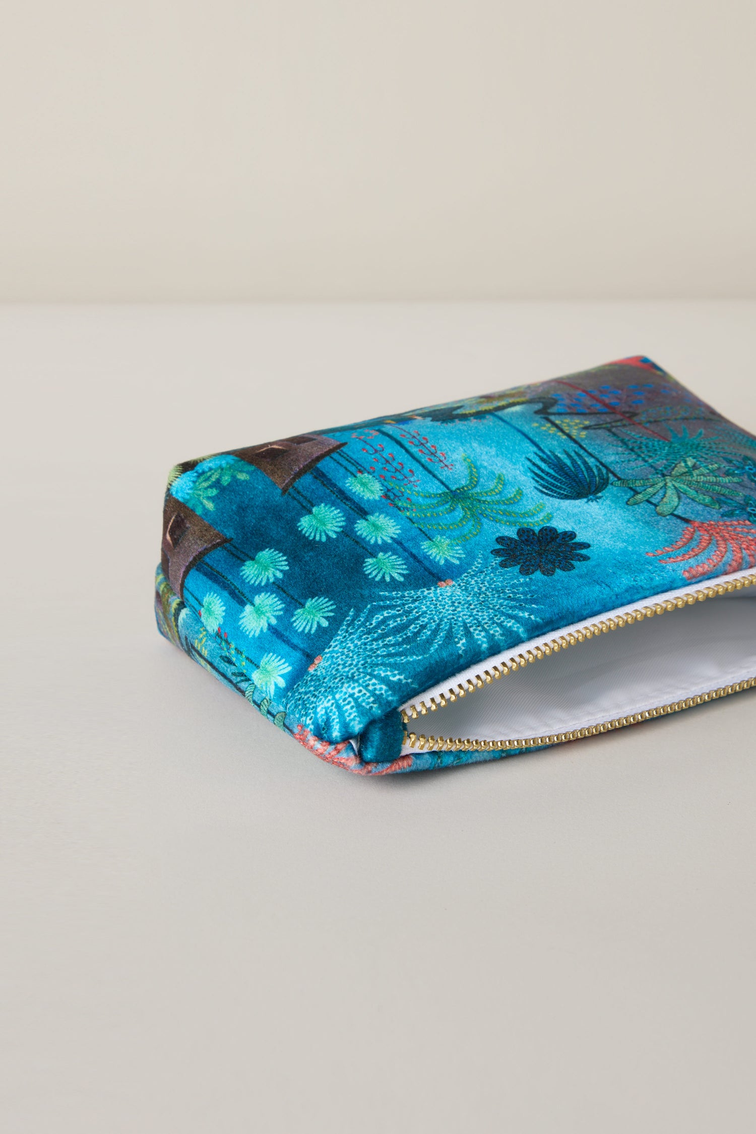 A Small Jungle Pathway Velvet Bag, similar to Daphne Stephenson's design, features vibrant floral and foliage patterns on a blue zippered pouch resting on a light surface.