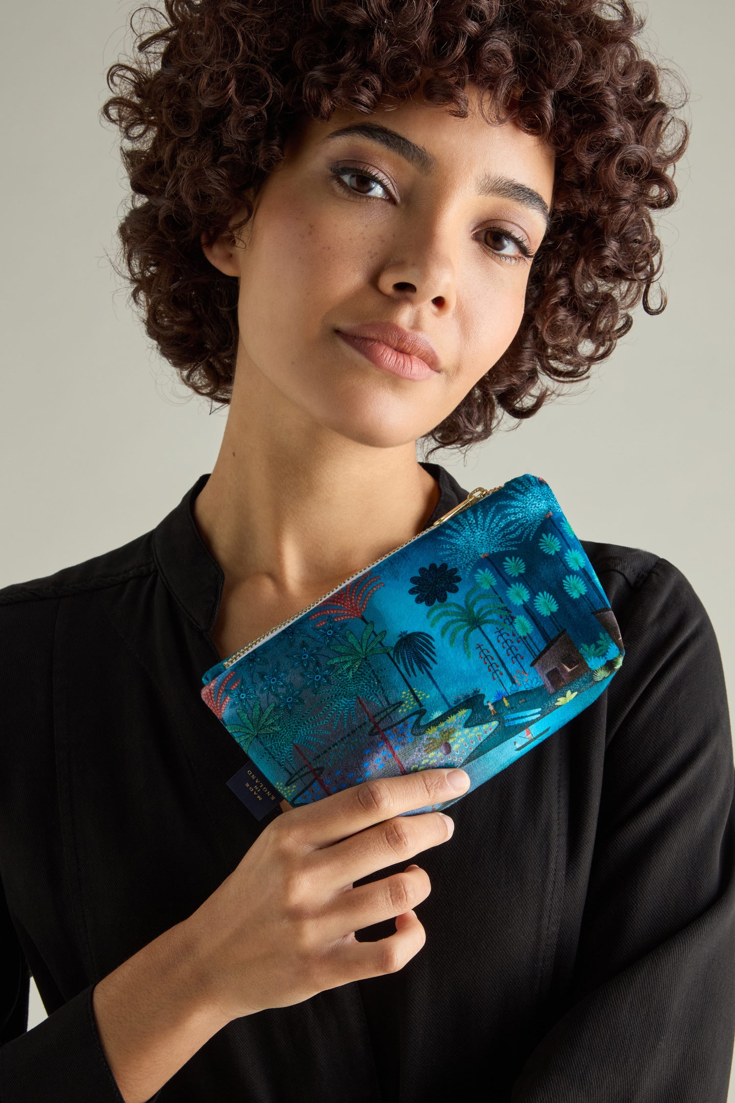 A person with curly hair holds a Small Jungle Pathway Velvet Bag with a tropical design, wearing a black shirt against a neutral background.