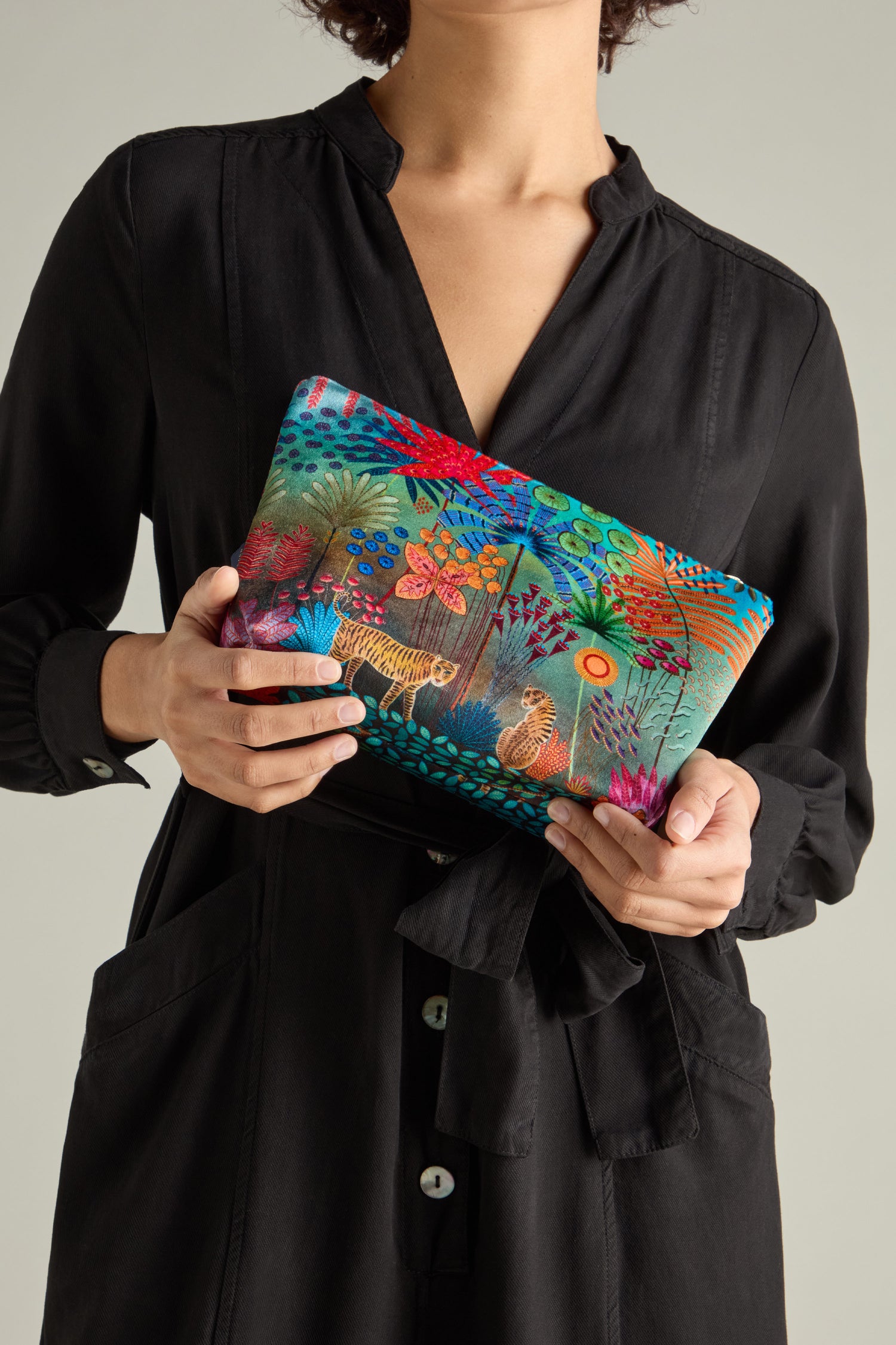 A person in a black dress holds the Large Tale of Two Tigers Velvet Bag by Daphne Stephenson, featuring a vibrant jungle-themed design.