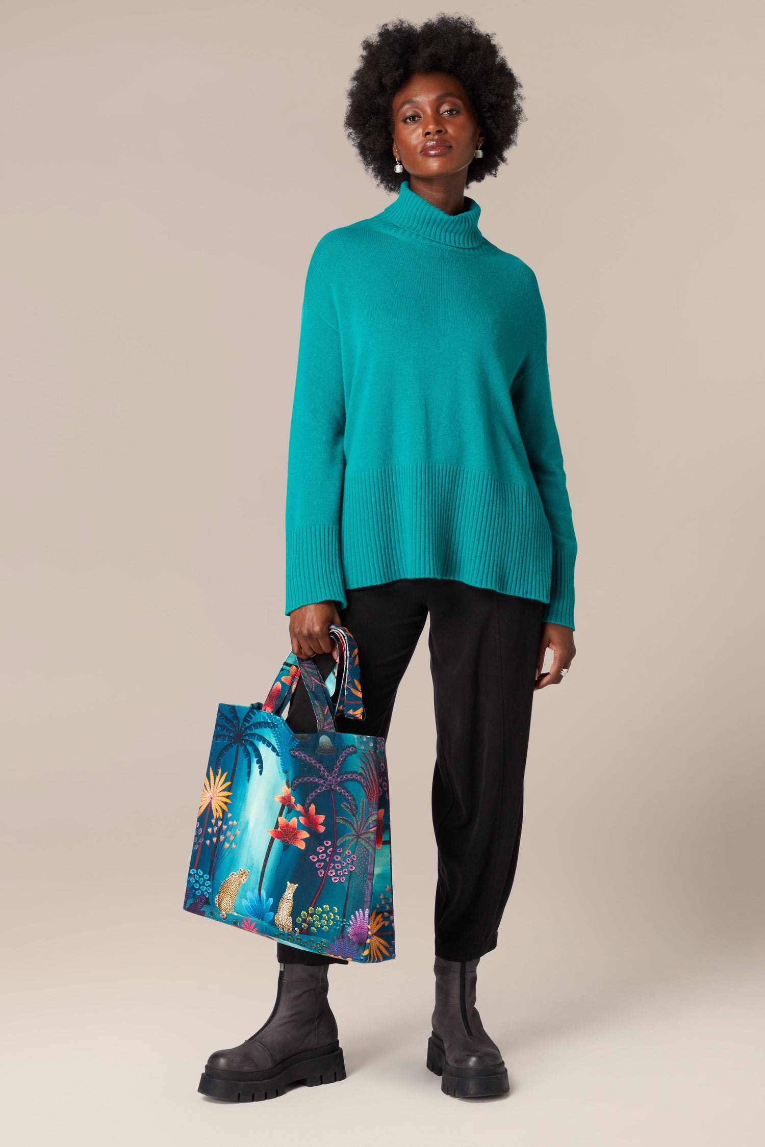 A woman wearing a turquoise sweater and black pants is holding a Twilight Jungle tote bag.