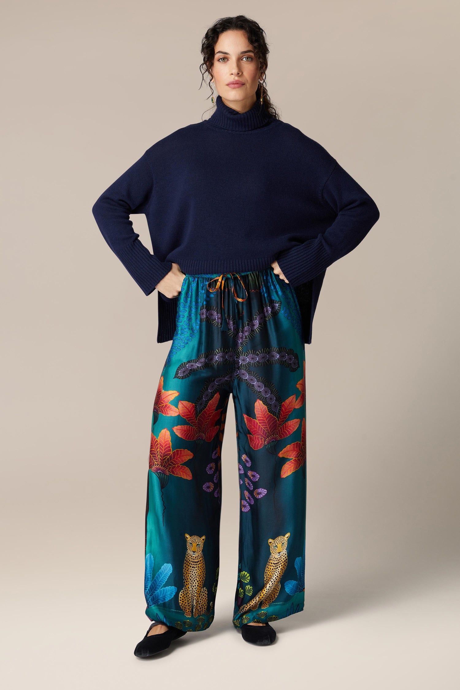 A woman wearing Twilight Jungle Trousers, wide-leg trousers with an exotic tropical print, paired with a blue turtleneck.