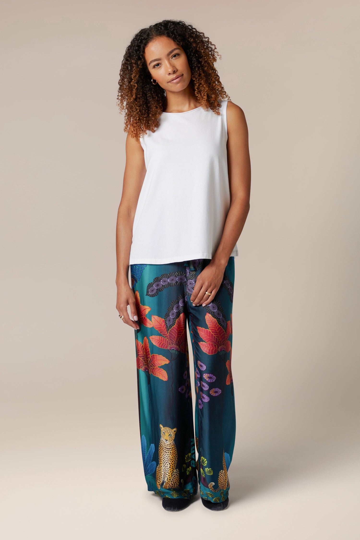 A woman in a white top adorned with a vibrant floral print on her Twilight Jungle Trousers, exuding an exotic style that transports you to tropical scenes.