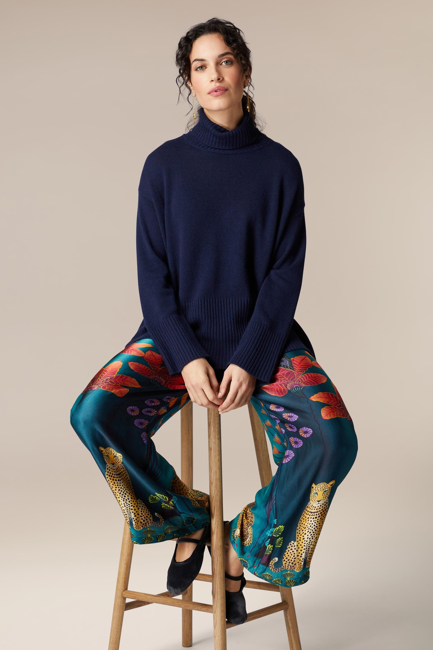 A woman is sitting on a stool in Twilight Jungle Trousers and a blue sweater.