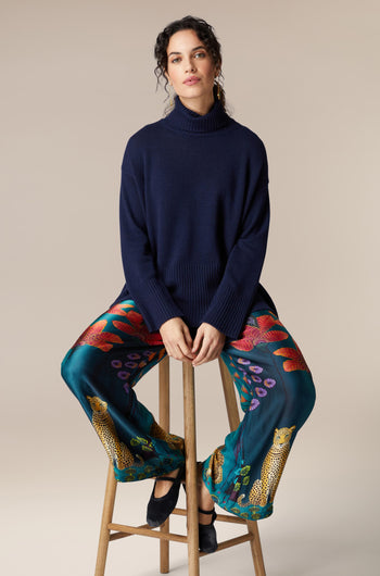 A woman is sitting on a stool in Twilight Jungle Trousers and a blue sweater.