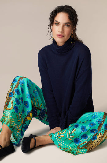 A woman is sitting on the floor in a blue sweater and green pants, showcasing her Leopard in Moonlight Trousers in an exotic style.