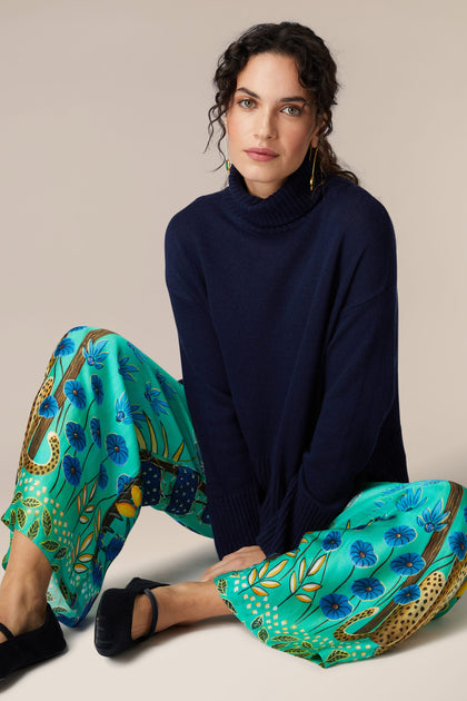 A woman is sitting on the floor in a blue sweater and green pants, showcasing her Leopard in Moonlight Trousers in an exotic style.