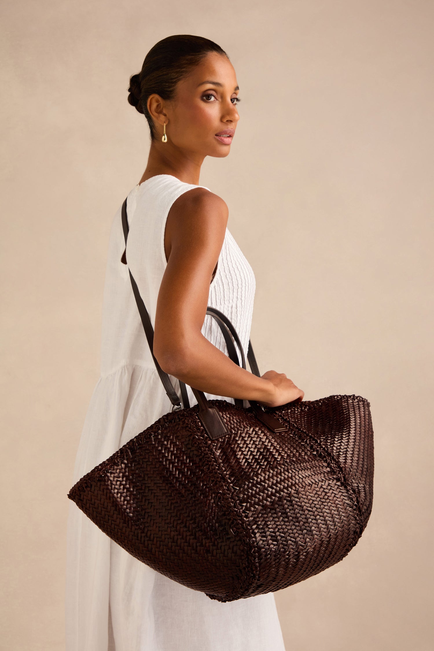 A woman in a sleeveless white dress holds the Teresa Woven Leather Bag—a large, dark brown handwoven leather accessory—over her shoulder, standing against a neutral background for an elegant display of craftsmanship.