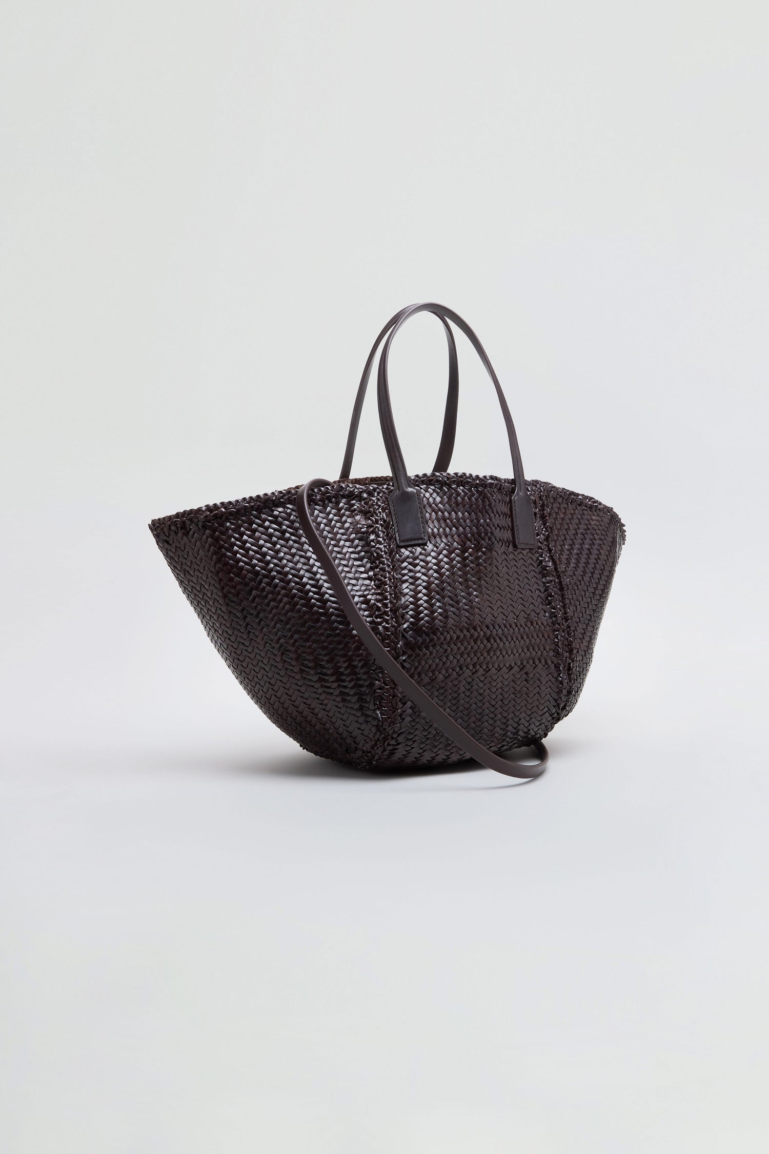 The Teresa Woven Leather Bag, crafted using traditional weaving techniques in dark brown leather, features two top handles and a long shoulder strap, and is displayed on a plain light background.