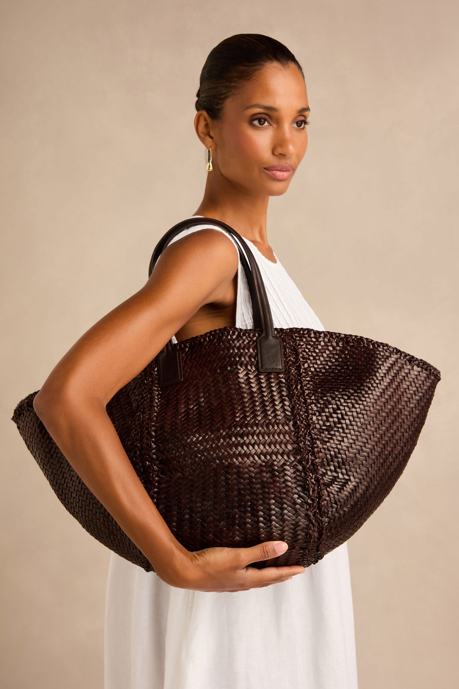 A woman in a white sleeveless dress holds the Teresa Woven Leather Bag—a large, dark brown tote with black handles, expertly crafted using traditional weaving techniques—against a neutral background.