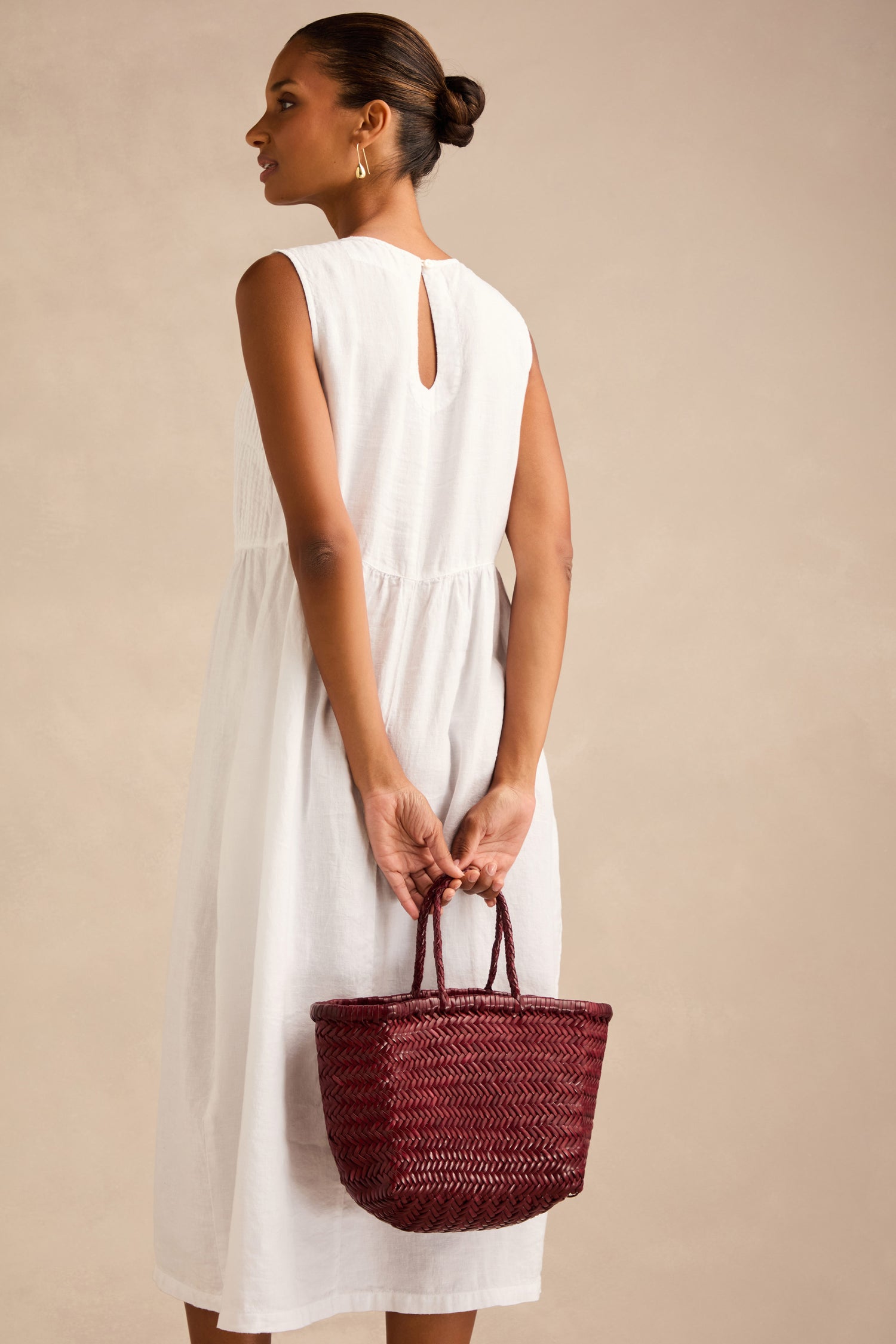 A woman in a sleeveless white dress stands facing away, holding a maroon Small Triple Jump Woven Leather Bag behind her back against a neutral background.