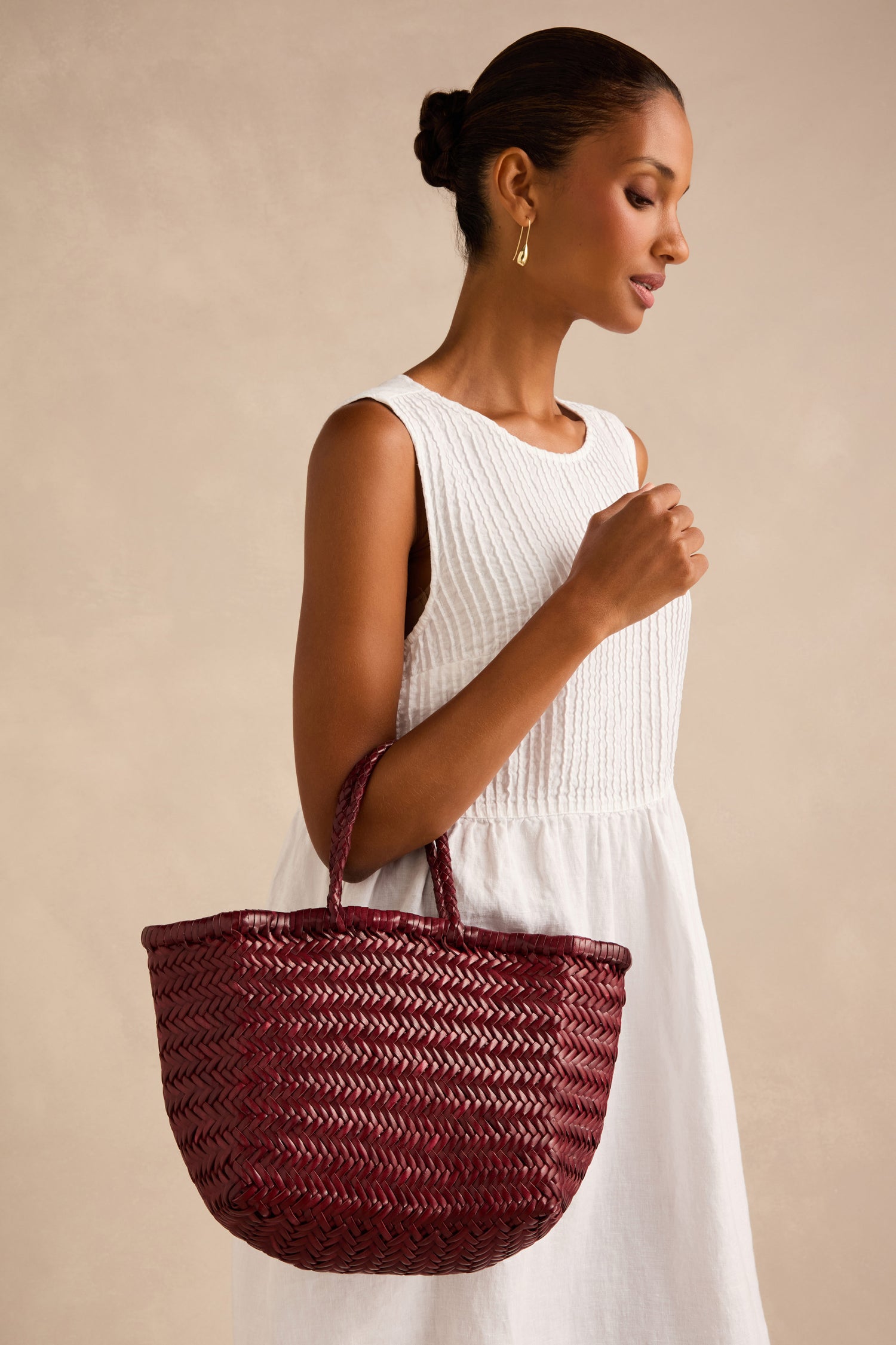 A woman with her hair in a bun and gold hoop earrings stands against a beige background, wearing a sleeveless white dress and holding the Dragon Diffusion Small Triple Jump Woven Leather Bag in maroon.