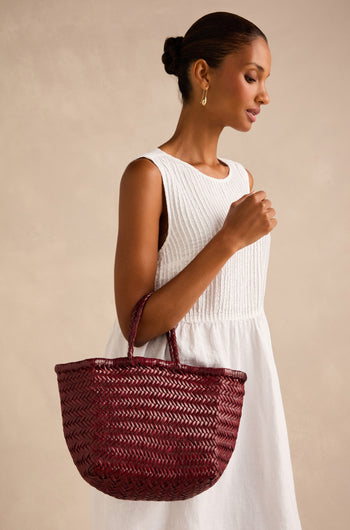 A woman with her hair in a bun and gold hoop earrings stands against a beige background, wearing a sleeveless white dress and holding the Dragon Diffusion Small Triple Jump Woven Leather Bag in maroon.