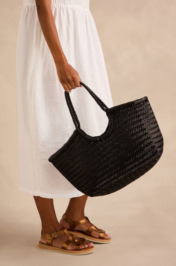 Person in a white dress and brown sandals holding a large Nantucket Woven Black Leather Bag, crafted from handwoven buffalo calf leather, against a neutral background.