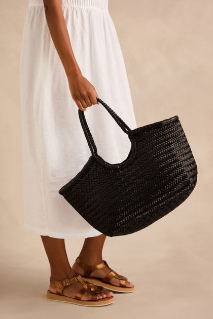 Person in a white dress and brown sandals holding a large Nantucket Woven Black Leather Bag, crafted from handwoven buffalo calf leather, against a neutral background.