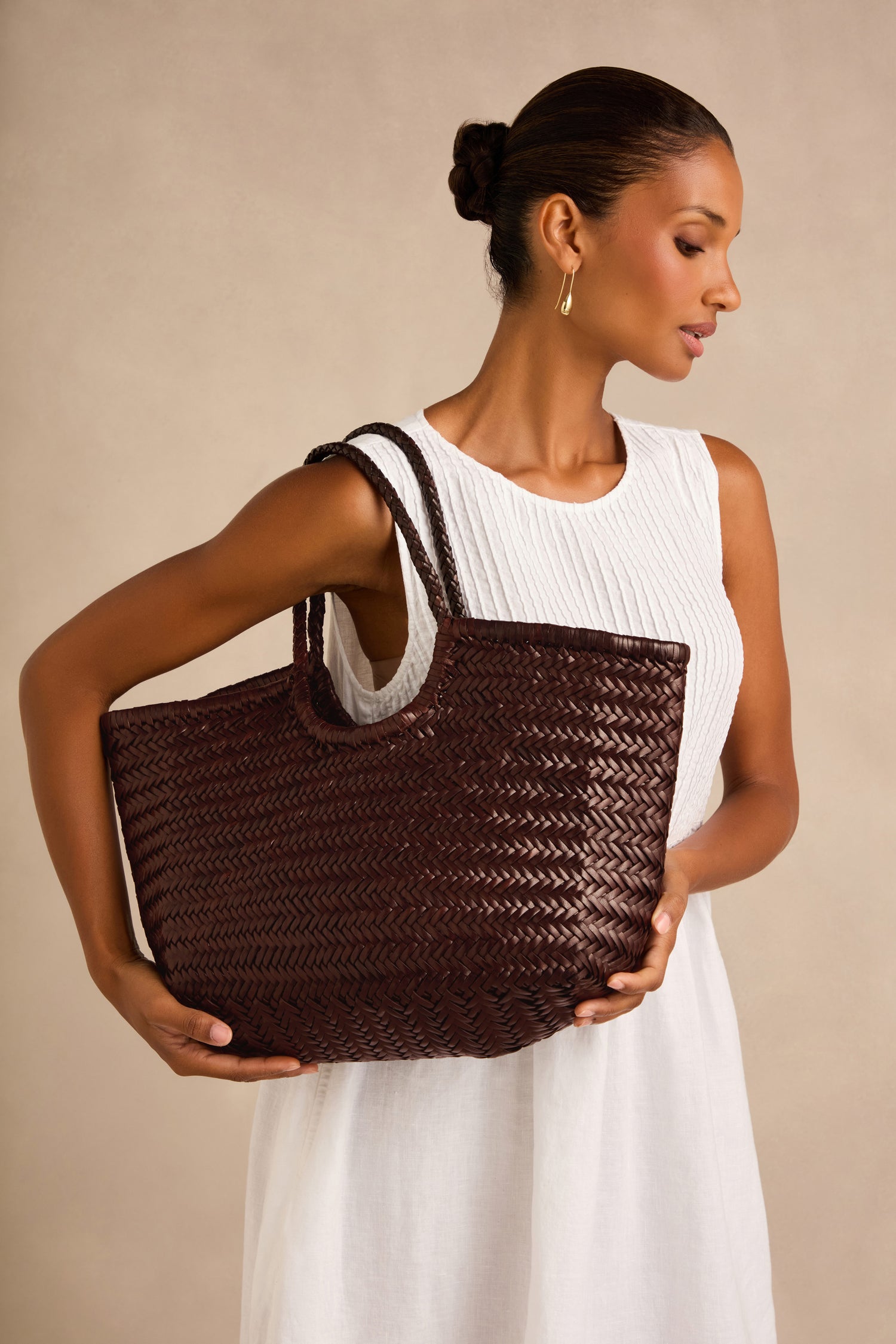 Woman in a sleeveless white dress holding a large Nantucket Woven Dark Brown Leather Bag, standing against a plain beige background.