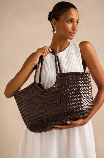 A woman in a sleeveless white dress holds a large Nantucket Woven Dark Brown Leather Bag with two handles against a neutral background.