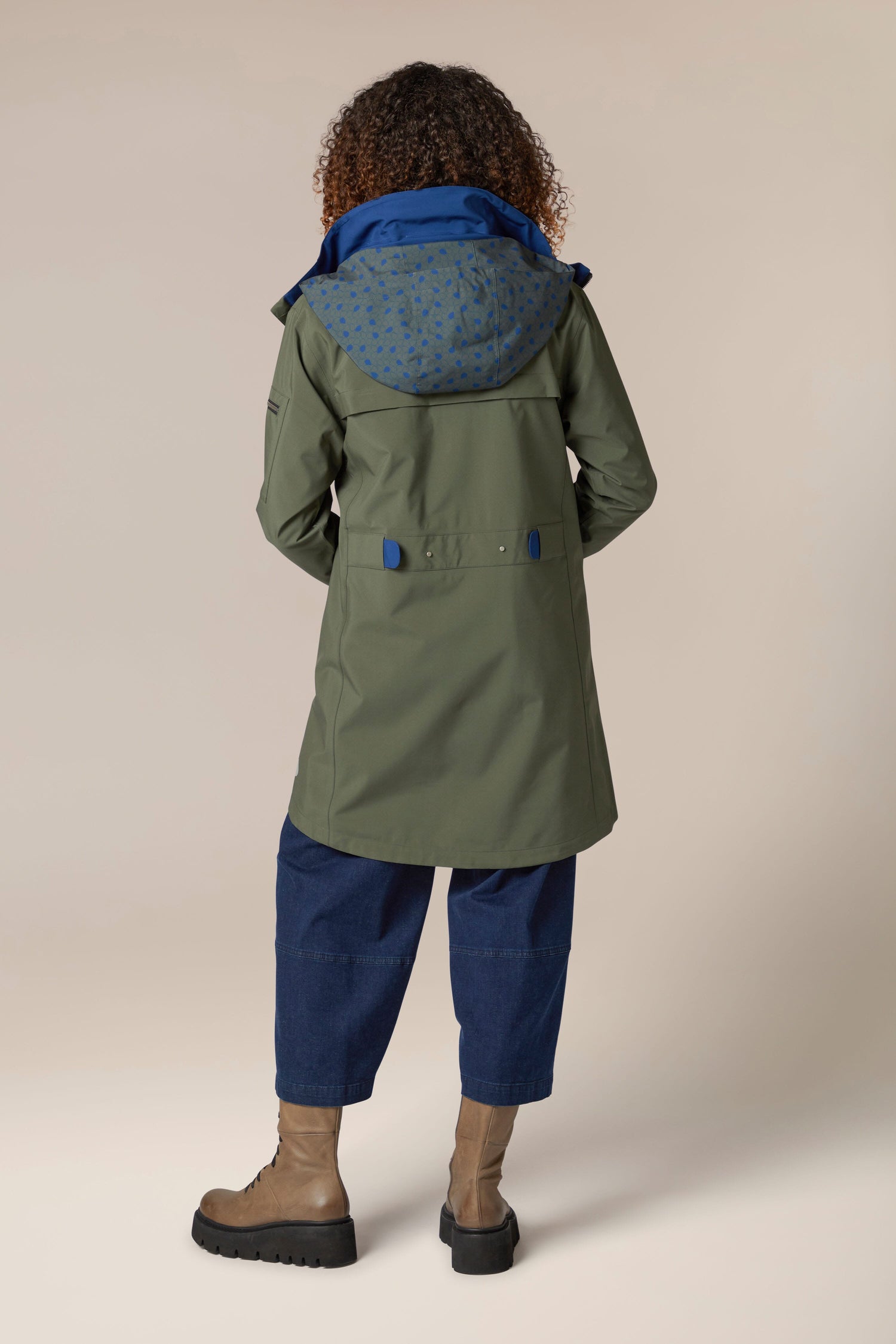 The back view of a woman wearing a 5 Pocket Hooded Rain Jacket from Regn, with adjustable wrists and blue boots.