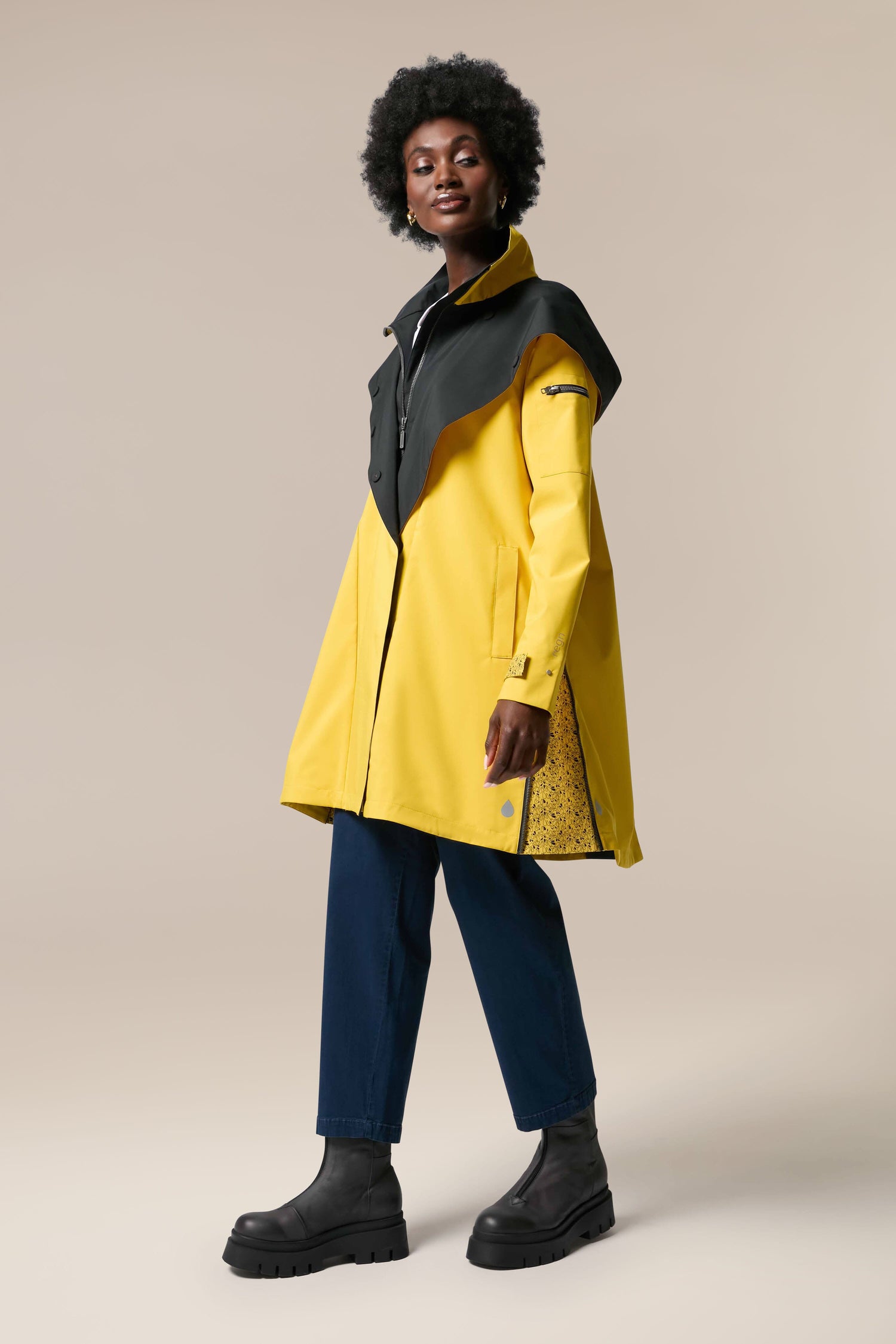 A woman wearing a yellow raincoat, the Denise Wide Collar Rain Jacket from Scandi brand Regn, paired with black boots.