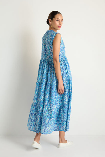 A woman models the Floral Tiered Dress, a sleeveless blue and white floral piece, paired with white sneakers as she stands against a plain white background, glancing over her shoulder.