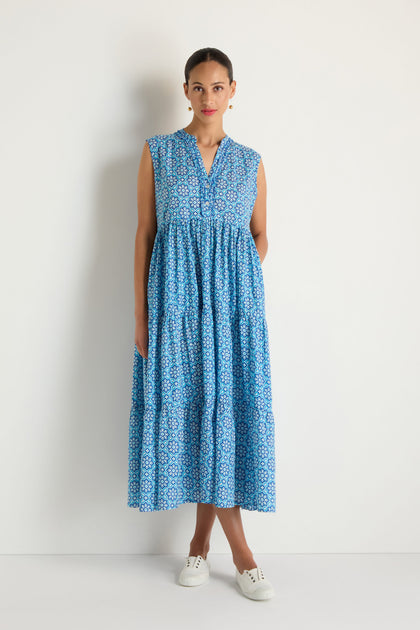 A woman stands against a plain white background wearing the Floral Tiered Dress, a sleeveless blue and white floral design, paired with white sneakers.