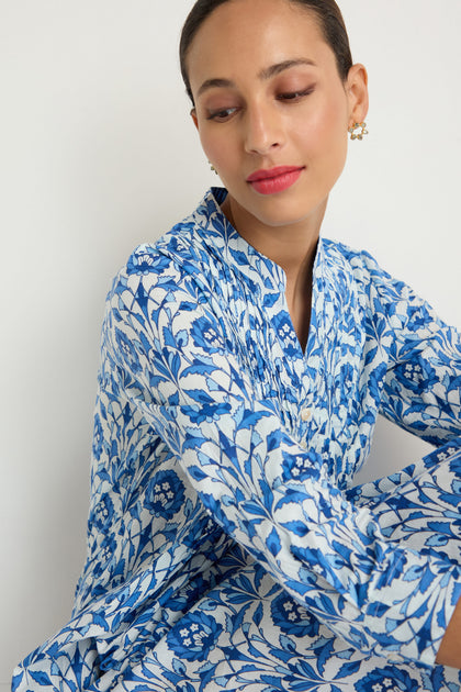 A woman with pulled-back hair wears the Block Print Pleat Front Dress, a blue and white floral cotton piece, sitting against a white background and looking down with a neutral expression.