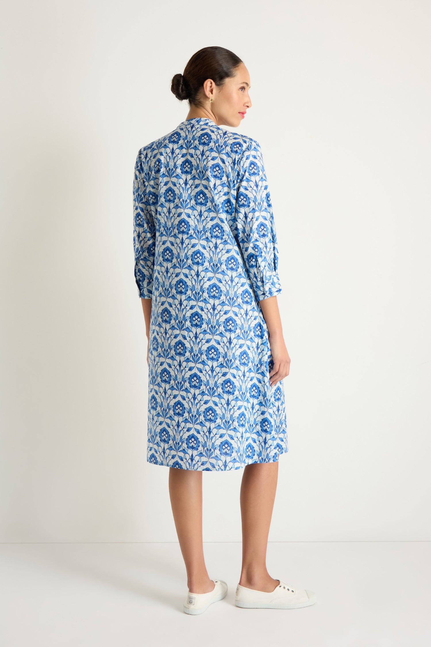 A woman faces away, wearing the Block Print Pleat Front Dress in blue and white with three-quarter sleeves and white sneakers, set against a plain white background.