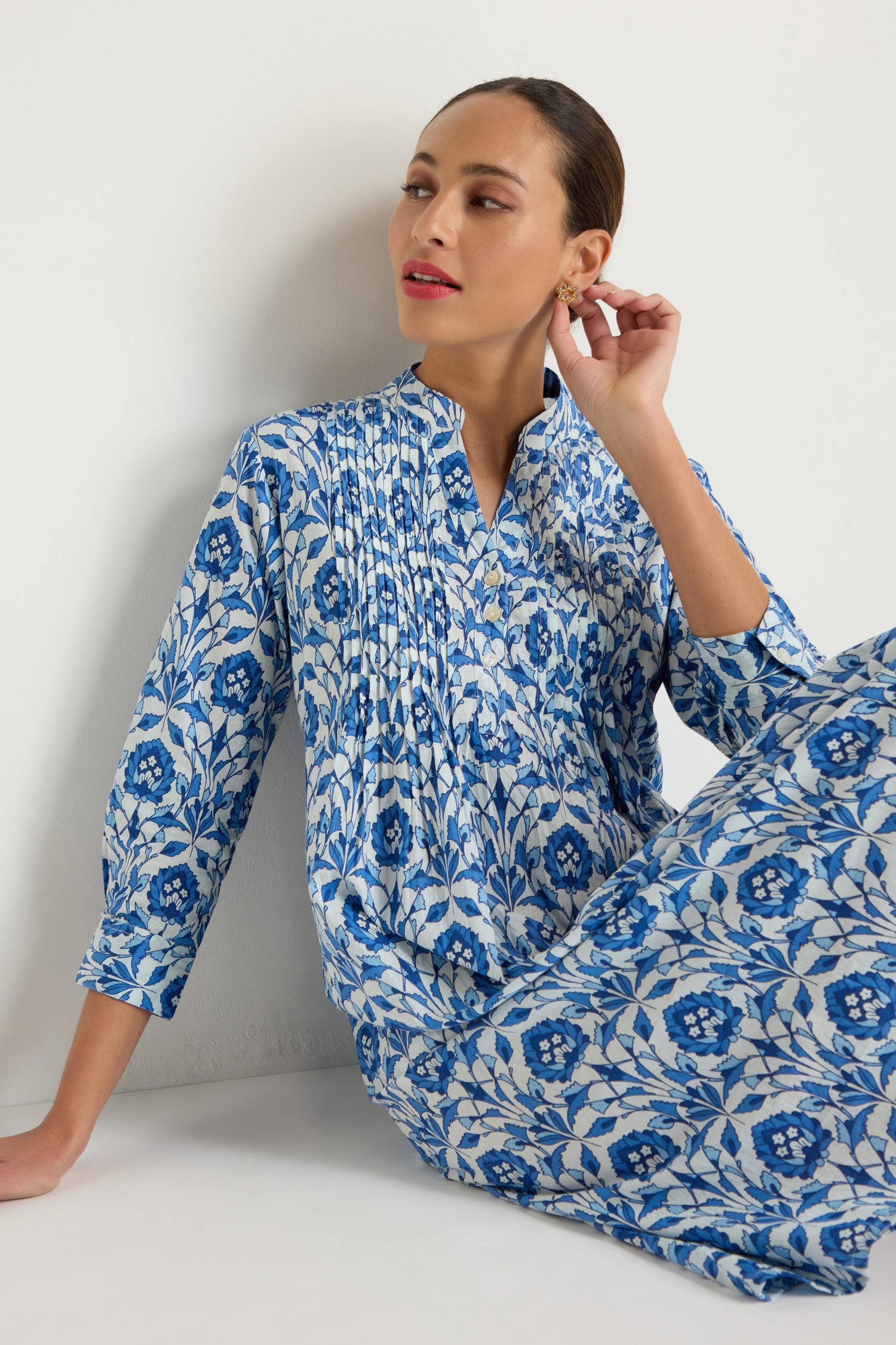 A woman wearing the Block Print Pleat Front Dress, featuring blue and white floral cotton, sits against a white wall, gazing to the side while gently touching her earring.