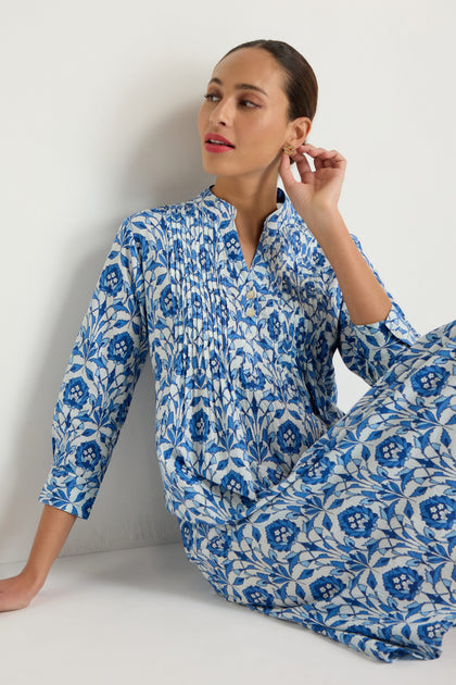 A woman wearing the Block Print Pleat Front Dress, featuring blue and white floral cotton, sits against a white wall, gazing to the side while gently touching her earring.
