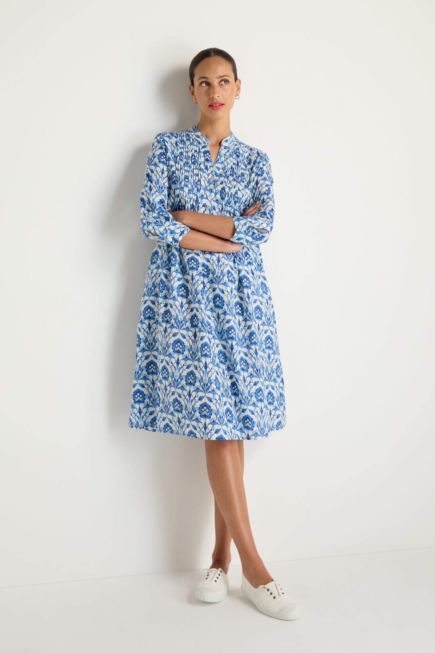 A woman stands against a white wall wearing the Block Print Pleat Front Dress with three-quarter sleeves and white sneakers, arms crossed and looking slightly to the side.