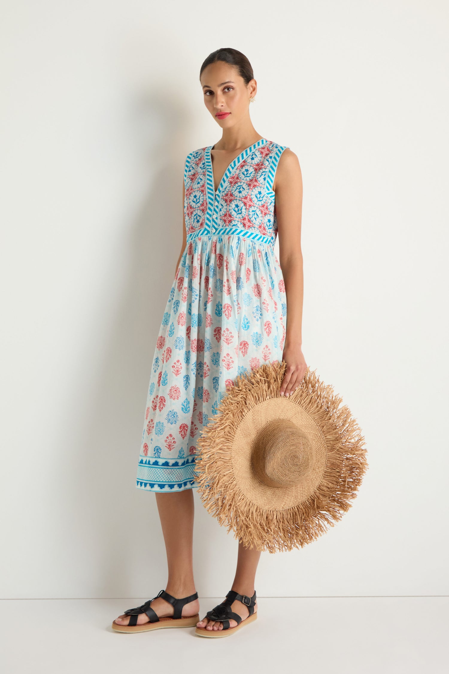 Woman against a white wall wears the Chevron Flared Dress in blue and red floral cotton block print, paired with black sandals and holding a large straw hat.