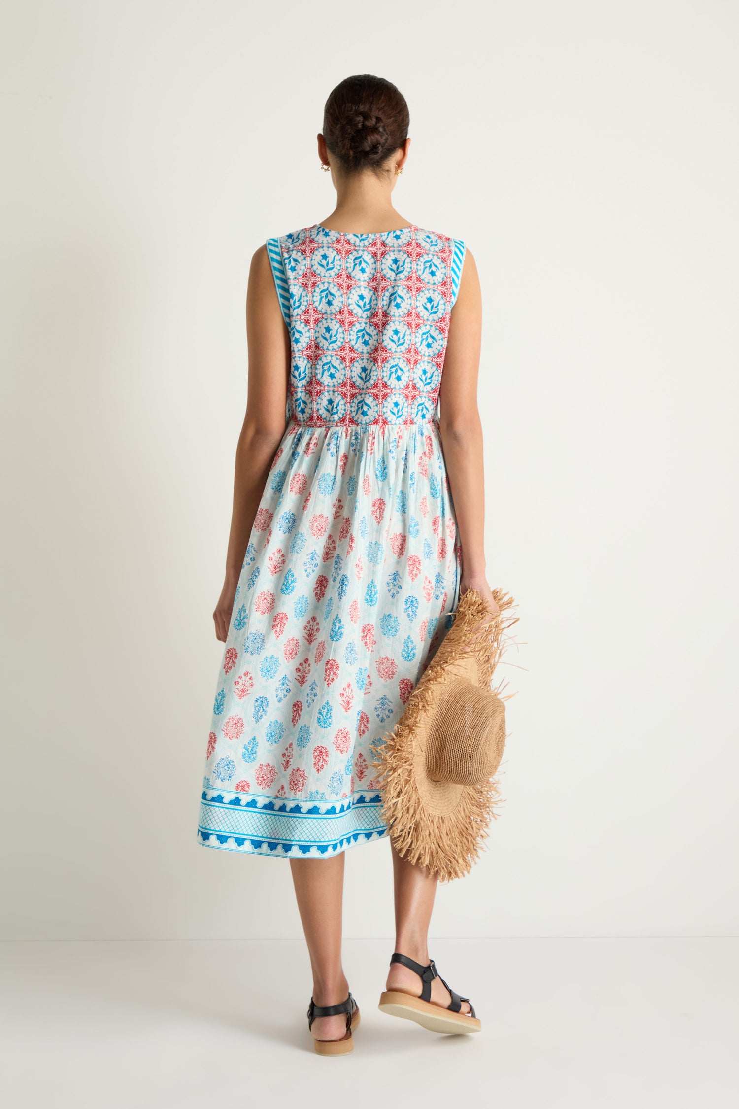 A woman faces away, wearing the Chevron Flared Dress—a sleeveless midi dress with blue and red cotton block prints—paired with black sandals and holding a straw hat against a plain background.