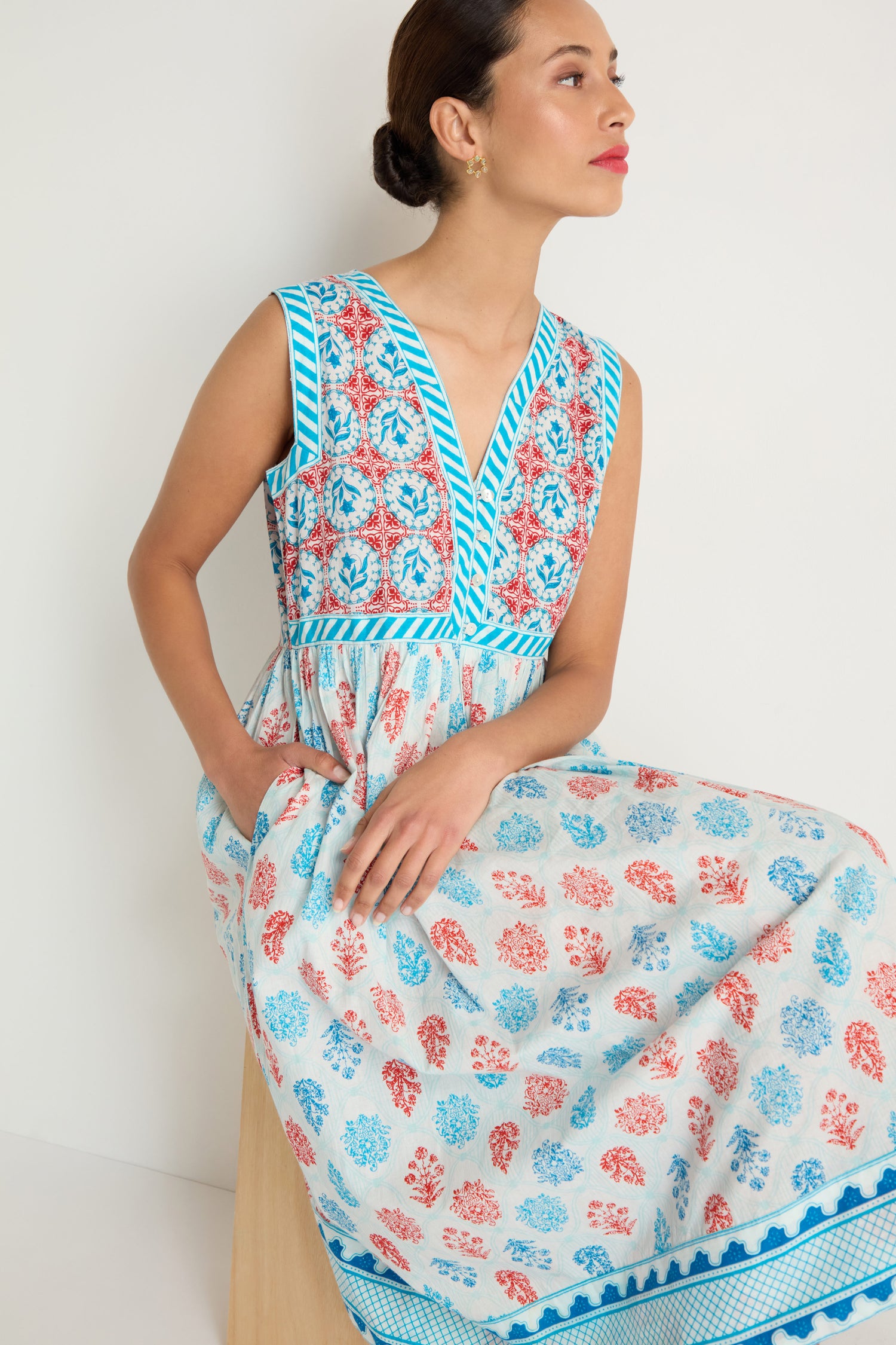 A woman sits on a wooden stool wearing the Chevron Flared Dress, a sleeveless midi with blue and red floral patterns and blue trim, crafted from soft cotton block print on a white background.